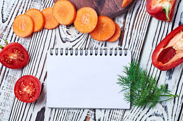 Different natural fresh ripe cutted vegetables near blank paper notepad lies on old white rustic wooden table on kitchen. Healthy food concept. Space for text. Top view - Photo, image