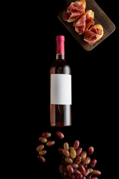 top view of rose wine bottle with blank white label near ripe grape and sliced prosciutto on baguette isolated on black - Foto, Imagem