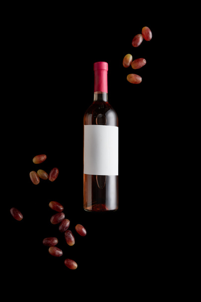 top view of rose wine bottle with blank white label near ripe grape isolated on black - Foto, afbeelding