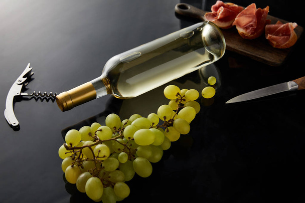 bottle with white wine near grape, knife, corkscrew and sliced prosciutto on baguette on black background - Photo, Image