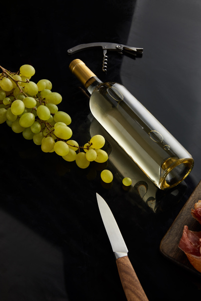 bottle with white wine near grape, knife and corkscrew on black surface - Foto, afbeelding
