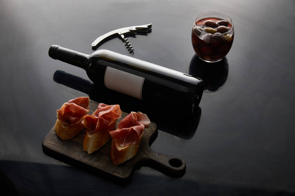 wine bottle with blank label near sliced prosciutto on baguette, corkscrew and olives on black background - Photo, Image