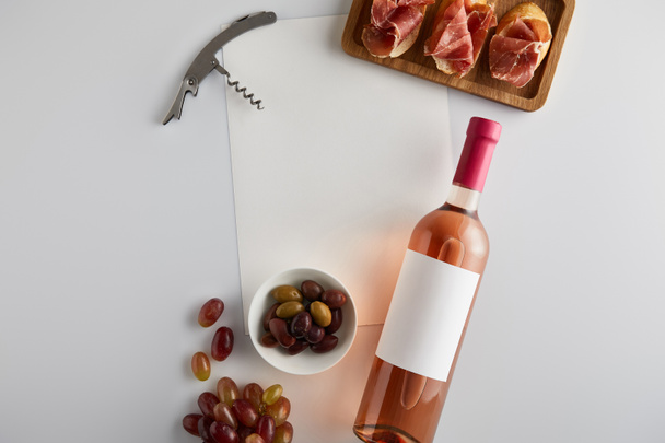 top view of bottle with rose wine near grape, corkscrew, olives, blank paper and sliced prosciutto on baguette on white background - Zdjęcie, obraz