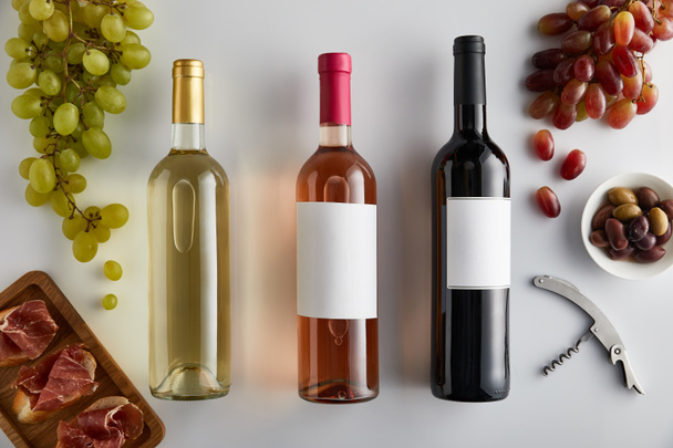 top view of bottles with white, rose and red wine near grape, corkscrew, olives and sliced prosciutto on baguette on white background - Photo, Image