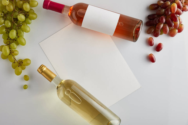 top view of bottles with white and rose wine near grape and blank paper on white background - Foto, afbeelding