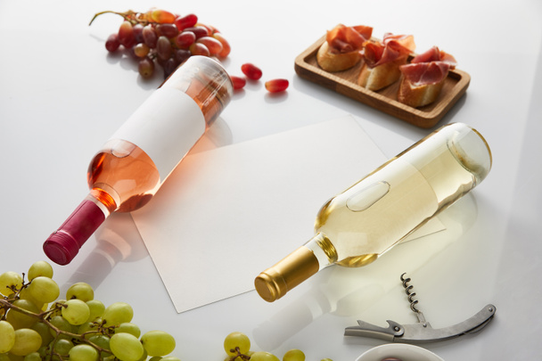 bottles with white, rose wine near grape, blank paper, corkscrew and sliced prosciutto on baguette on white background - Photo, Image