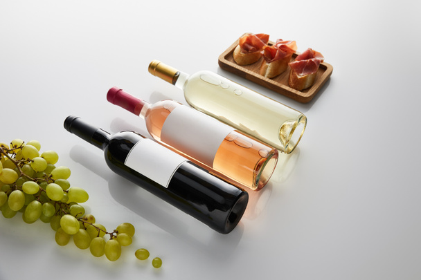bottles with white, rose and red wine near grape and sliced prosciutto on baguette on white background - 写真・画像