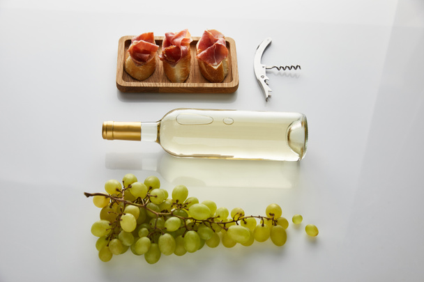 flat lay with bottle with white wine near grape, corkscrew and sliced prosciutto on baguette on white background - Photo, Image