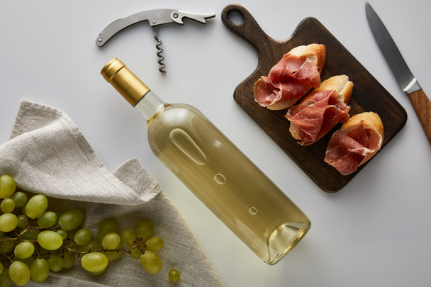 felső nézet palack fehér borral közel szőlő, dugóhúzó, kés és szeletelt prosciutto fehér alapon - Fotó, kép