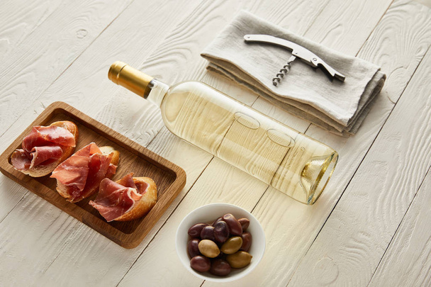bottle with white wine near prosciutto on baguette, olives and corkscrew on napkin on white wooden surface - Photo, Image