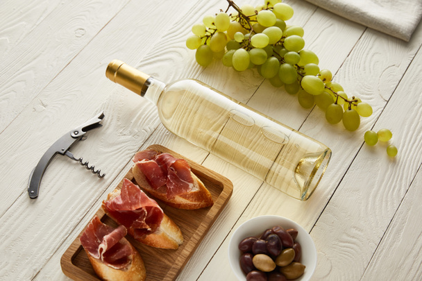 bottle with white wine near grape, prosciutto on baguette, olives and corkscrew on white wooden surface - Фото, изображение