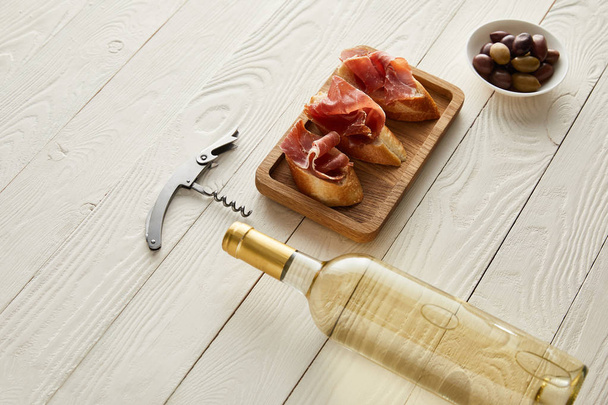 bottle with white wine near prosciutto on baguette, olives and corkscrew on white wooden surface - Photo, Image