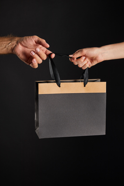 przycięty widok kobiety i mężczyzny trzymających papierową torbę na zakupy odizolowany na czarny, czarny piątek koncepcja - Zdjęcie, obraz