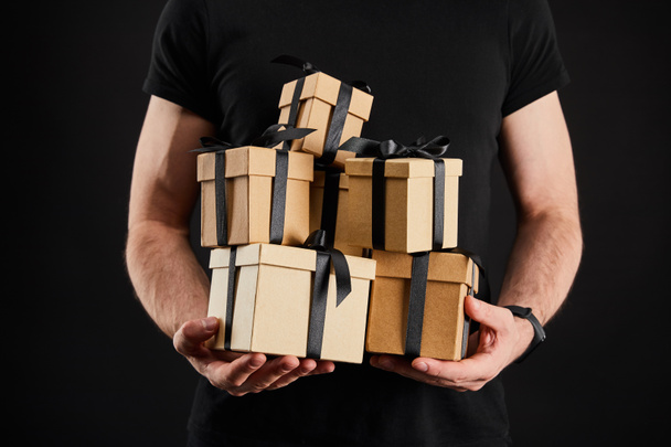 partial view of man holding cardboard gift boxes with ribbons isolated on black, black Friday concept - Valokuva, kuva