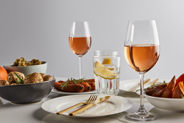 glazen met rozenwijn en citroenwater, gebakken aardappelen en wortelen, witte borden en bestek op marmeren tafel geïsoleerd op grijs - Foto, afbeelding