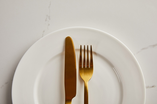 Altın bıçak ve çatalın üst görüntüsü mermer masada temiz beyaz tabak üzerinde - Fotoğraf, Görsel