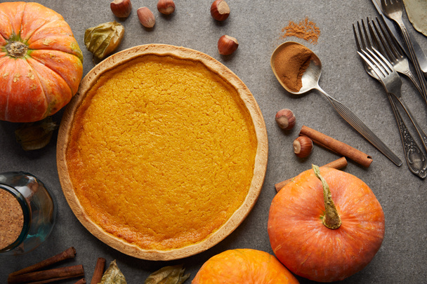 top view of delicious pumpkin pie near whole pumpkins, cutlery and hazelnuts on grey stone surface - Photo, image