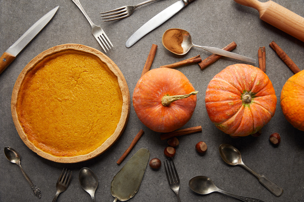 top view of tasty pumpkin pie near whole ripe pumpkins, cutlery, cinnamon sticks and hazelnuts on grey stone surface - Photo, Image