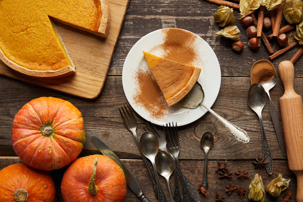 top view of tasty pumpkin pie on plate with spatula near whole pumpkins and cutlery on dark wooden surface - Photo, Image