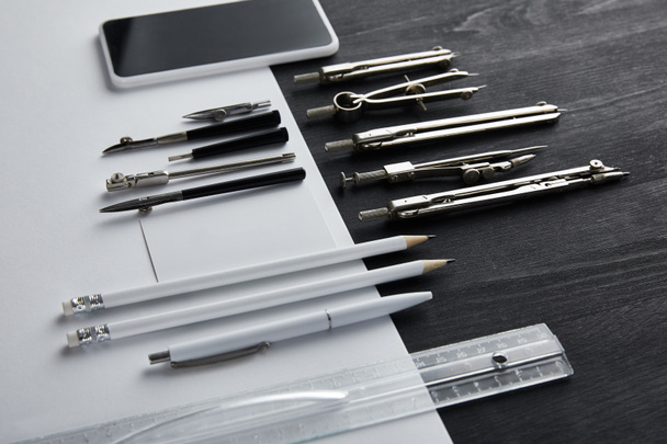 bílý papír s kopírovacím prostorem a kompasy, pravítko, tužky, smartphone, pero na černém a dřevěném stole  - Fotografie, Obrázek