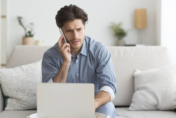 Guy έχοντας δυσάρεστη τηλεφωνική συνομιλία εργασίας στο laptop στο σπίτι - Φωτογραφία, εικόνα