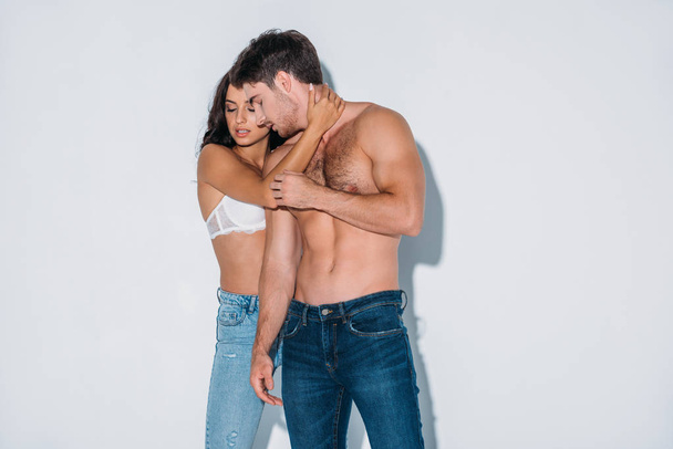 sexy girl in blue jeans and white bra embracing shirtless boyfriend on grey background - Фото, изображение