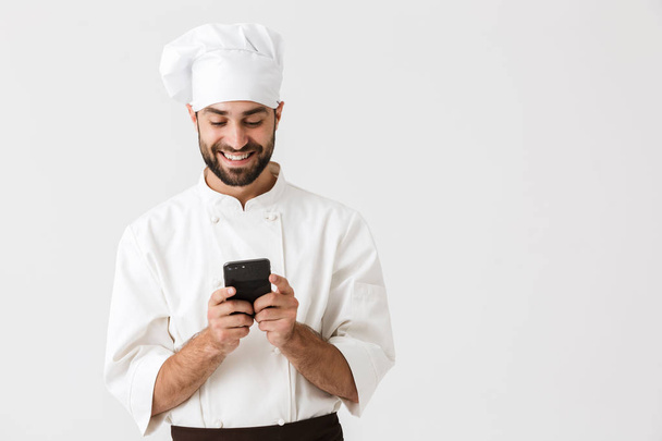 Cheerful smiling young chef posing isolated over white wall background in uniform using mobile phone. - Foto, Imagen
