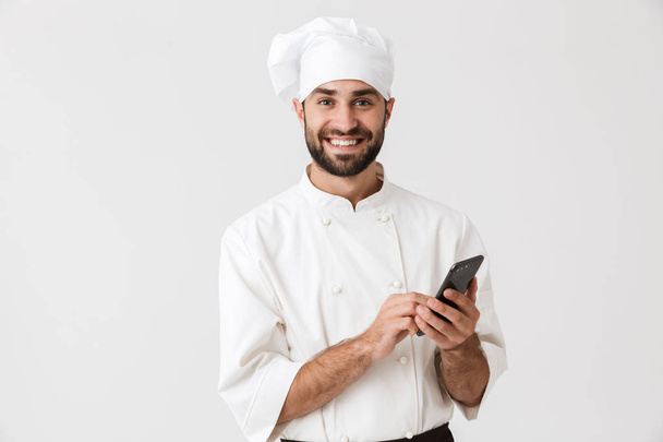 Cheerful smiling young chef posing isolated over white wall background in uniform using mobile phone. - Fotó, kép
