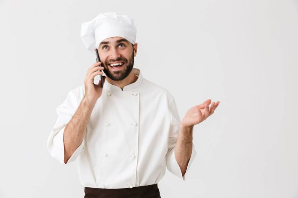 Emotional happy young chef posing isolated over white wall background in uniform talking by mobile phone. - Fotoğraf, Görsel