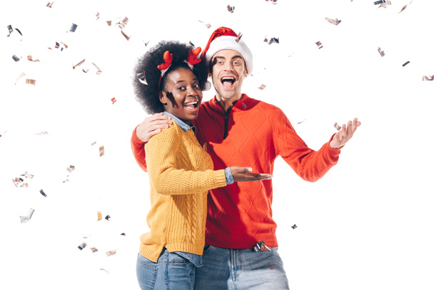 glückliches gemischtrassiges Paar mit Weihnachtsmann und Hirschhörnern, das mit Konfetti feiert, isoliert auf weißem Grund - Foto, Bild