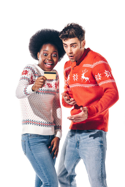 smiling multicultural couple in winter sweaters holding credit card, isolated on white - Foto, imagen