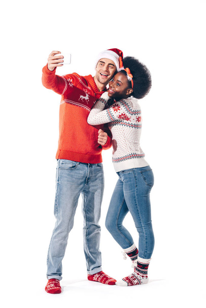 hermosa pareja interracial en sombrero de santa y cuernos de ciervo tomando selfie en el teléfono inteligente, aislado en blanco
 - Foto, Imagen