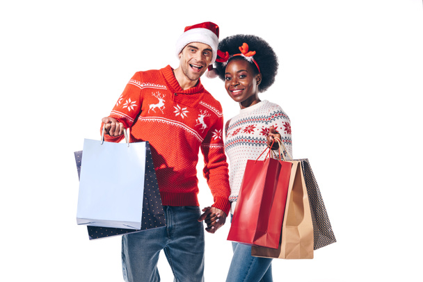 multicultural couple in santa hat and deer horns holding shopping bags, isolated on white - 写真・画像
