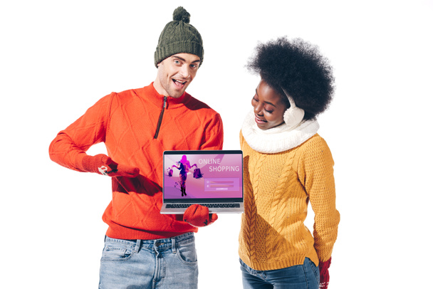 wielokulturowa para w zimowych swetrach, rękawiczkach i kapeluszu z laptopem na stronie internetowej sklepu internetowego, odizolowana na biało  - Zdjęcie, obraz