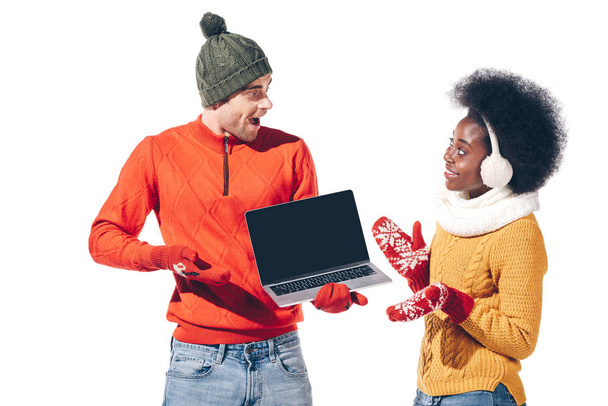 pareja multiétnica excitada en suéteres de invierno, guantes y sombrero que sostiene el ordenador portátil con pantalla en blanco, aislado en blanco
  - Foto, Imagen