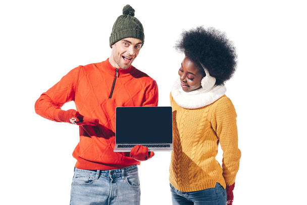 gyönyörű multikulturális pár téli pulóverben, kesztyű és kalap tartó laptop üres képernyőn, elszigetelt fehér  - Fotó, kép