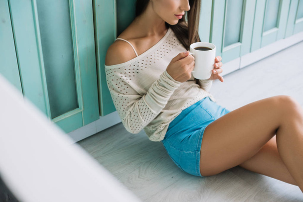 cropped view of woman holding cup of coffee and sitting on floor in kitchen - 写真・画像