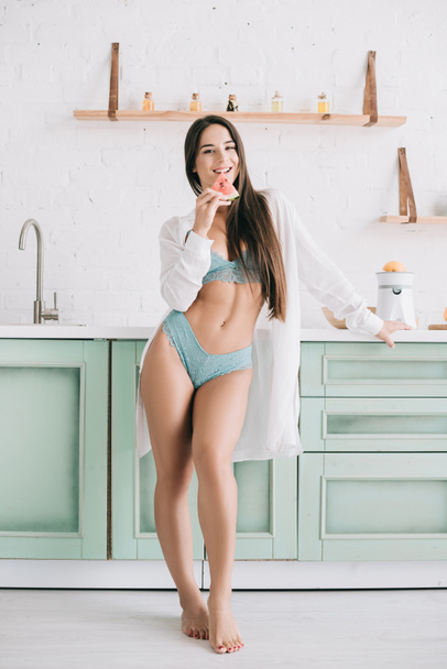 smiling sexy girl in lingerie and white shirt holding watermelon piece in kitchen - Photo, image