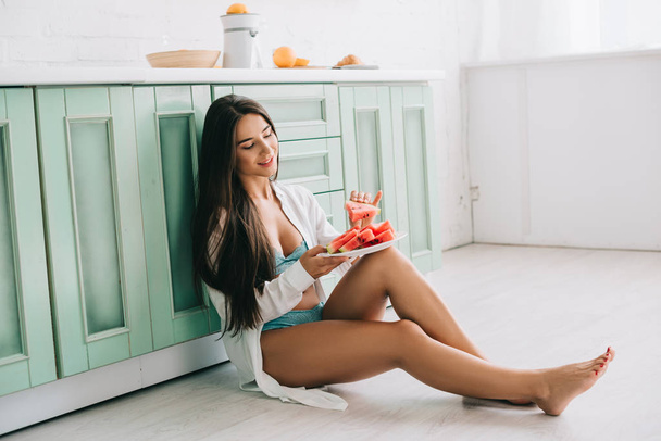 smiling woman in lingerie and white shirt eating watermelon on floor in kitchen - Photo, image