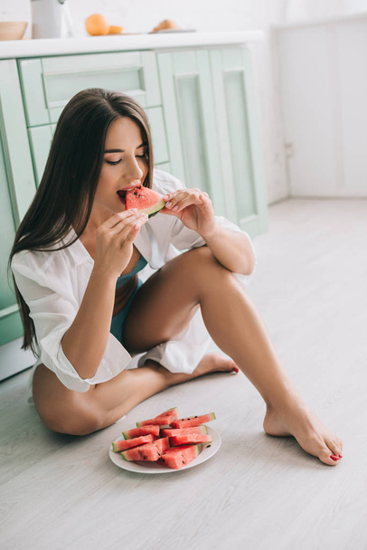 attractive woman in lingerie and white shirt eating watermelon on floor in kitchen - Photo, Image