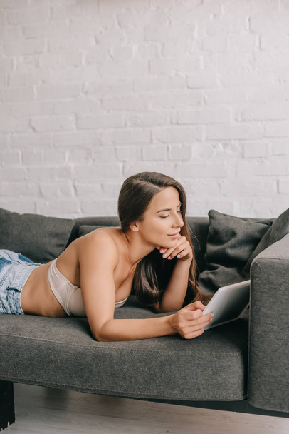 Beyaz sutyen ve kot pantolon giyen gülümseyen kadın oturma odasındaki kanepede dijital tablet kullanıyor. - Fotoğraf, Görsel