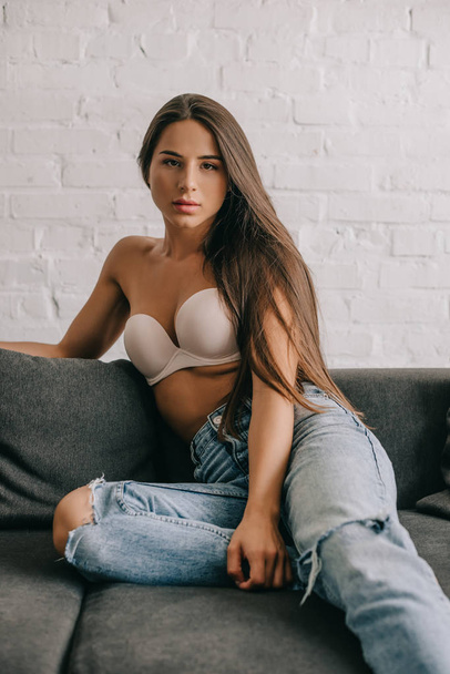 beautiful woman with long hair sitting on sofa in white bra and jeans - Фото, зображення