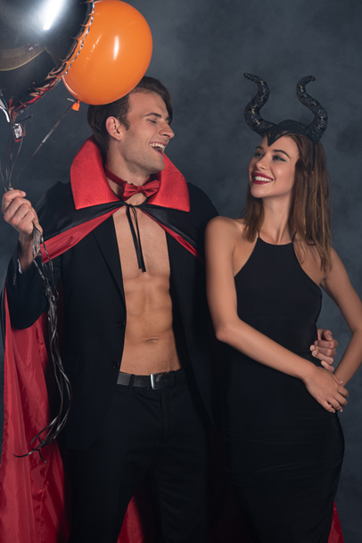 glücklicher Mann im Vampir-Halloween-Kostüm mit Luftballons in der Nähe von Mädchen mit Hörnern auf schwarz mit Rauch  - Foto, Bild