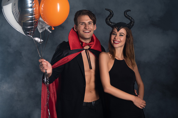 cheerful man in vampire halloween costume holding balloons near girl with horns on black with smoke  - Фото, изображение