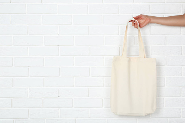 Female hand holding cotton eco bag on white brick wall backgroun - Zdjęcie, obraz