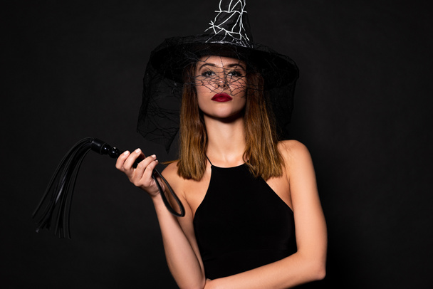 attractive woman in witch hat holding flogging whip isolated on black  - Foto, afbeelding