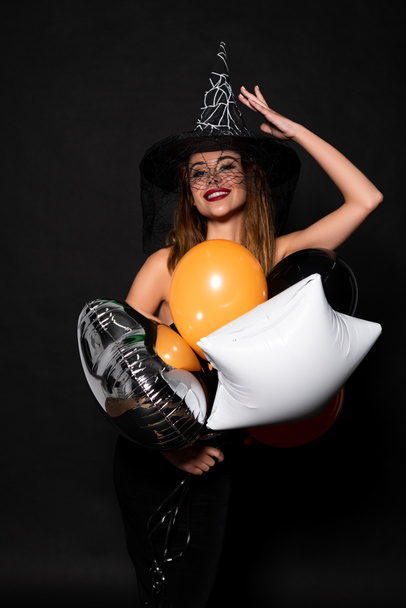 mujer feliz tocando sombrero de bruja cerca de globos en negro
  - Foto, Imagen