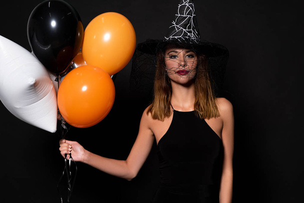 jovem mulher em chapéu de bruxa segurando balões isolados em preto
  - Foto, Imagem