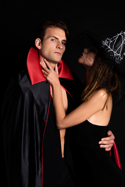 γυναίκα με καπέλο μάγισσας που αγγίζει το λαιμό του άνδρα με αποκριάτικη στολή που απομονώνεται σε μαύρο  - Φωτογραφία, εικόνα