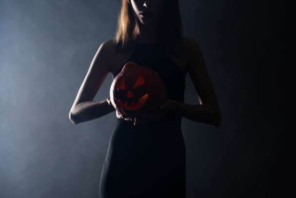 cropped view of woman with horns holding spooky pumpkin on black with smoke  - Foto, Bild